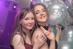 18th birthday party. 2 young women are smiling for their photo. Purple mobile disco light brightens up the background.