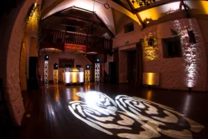 A wedding reception room with a mobile disco setup. Disco lights project roses onto the dance floor. Venue uplighting in gold and white light up the walls.