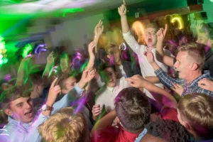 Crowd of students jump in the air at prom party. Coloured disco lights fill the background. A disc jockey plays music in the background. 