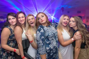 Six friends at pose for a selfie at prom party disco. Disc jockey plays music in the background. Laser beams and disco lights brighten up the room.