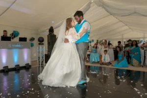 Bride and groom have romantic first dance at marquee wedding reception. The guests are watching. Wedding DJ plays their special song in the background. A starlit dance floor complements the venue uplighting in blue and pink.