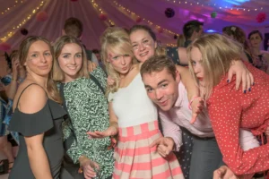 A group of friends at a marquee wedding reception smile for a picture. String lights and venue uplighting in pink and blue create a stunning background.