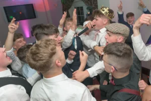 The prom king wears his crown and rocks the microphone over tracks played by a disc jockey at a school disco. He is surrounded by students celebrating their exam results. 